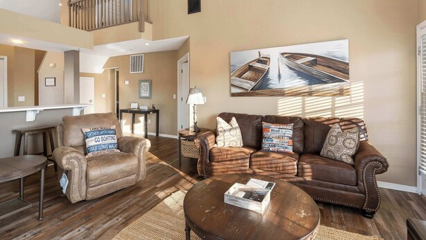 A spacious living room with a brown leather sofa, a brown leather armchair, and a coffee table. There is a painting of a boat on the wall, a staircase leading to an upper level, and a door with a window.