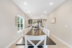 Une salle à manger avec une longue table, des chaises blanches, une plante en pot et une cuisine en arrière-plan.