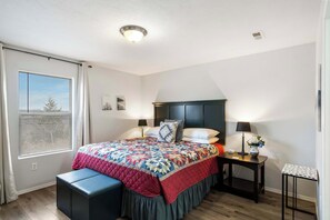 Un dormitorio con una cama, mesitas de noche, una ventana con persianas, una lámpara de noche y una pequeña mesa.