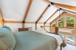 A tent-style room with a bed, wooden beams, a window, and a view of greenery.