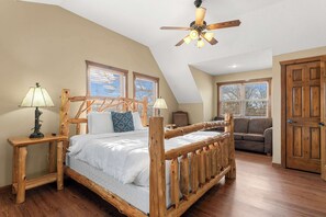 Un dormitorio con cama de madera, dos mesitas de noche, ventilador de techo y vistas al exterior a través de ventanas.