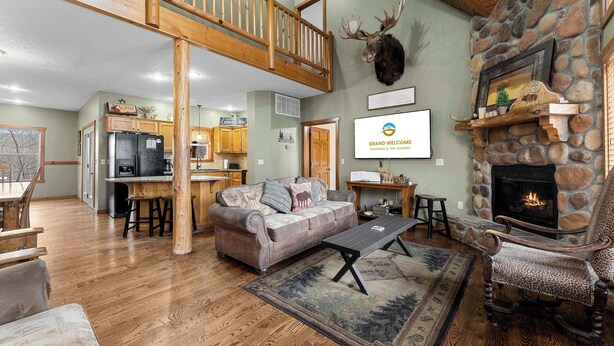 A spacious living area with a stone fireplace, a wooden staircase, and a kitchen in the background. - Royal Moose Lodge – A Grand Adventure for 20! (Branson)