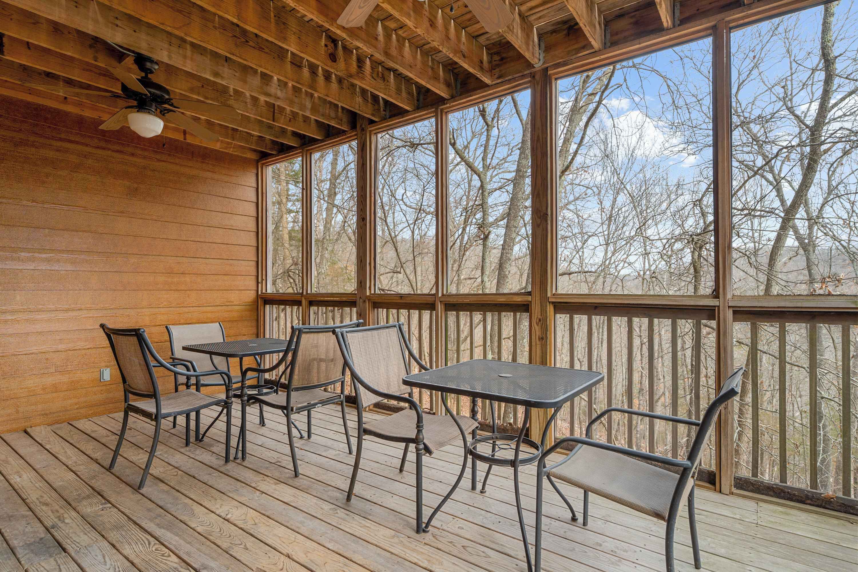 Une terrasse extérieure couverte, dotée d’un plancher en bois, de tables et de chaises en métal, et de grandes fenêtres offrant une vue sur les arbres et les montagnes.