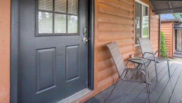 A log cabin with a dark front door, a window with blinds, a porch with two metal chairs, and a small green plant. - Cave Run Lodge - Near Lake - Playground & Fire Pit- Cabin #5 (Morehead)