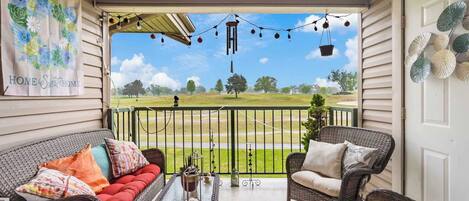 Eine überdachte Veranda mit Korbmöbeln, Blick auf einen Golfplatz und dekorative Lichterketten.