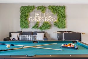 Una mesa de billar con bolas, una mesa de futbolín y una pared decorada con follaje verde y un cartel que dice 'LET THE GAMES BEGIN'.