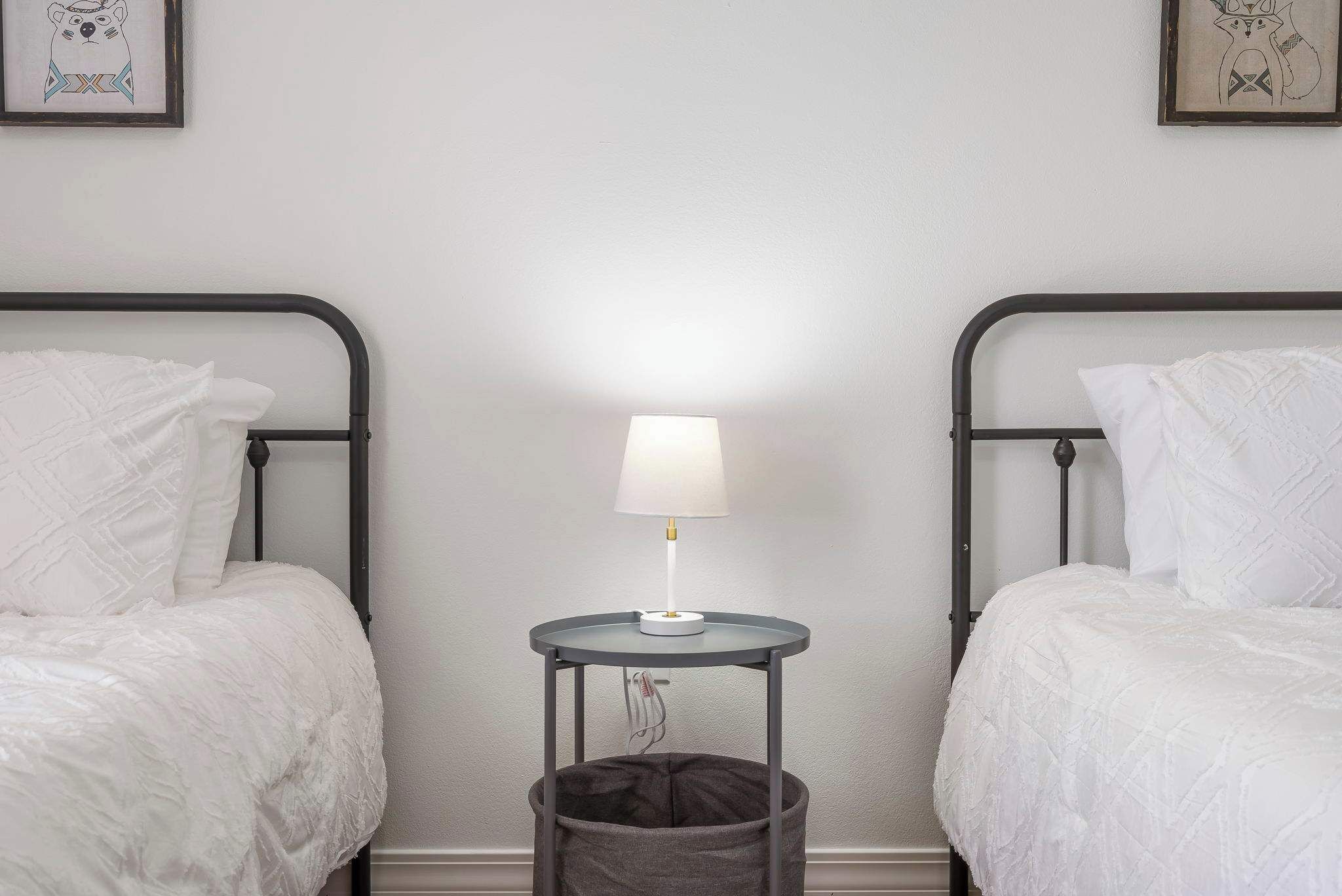 A bedroom with two beds, a bedside table with a lamp, and a waste bin.