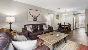 A modern living room with a leather sofa, a coffee table, and a dining area with a chandelier. - Urban Farmhouse | Refreshing Pool! (Branson)