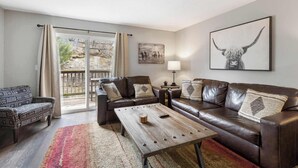 A living room with a brown leather sofa, a patterned armchair, a wooden coffee table, and a large window with curtains. - Urban Farmhouse | Refreshing Pool! (Branson)