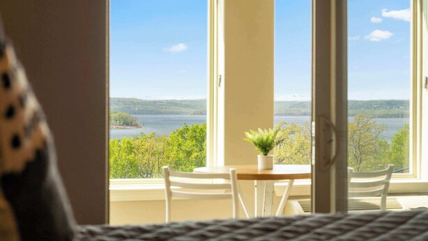A room with a large window offering a view of a lake and trees. There is a small table with a potted plant and two chairs. - It's a Good Life | Stunning Lake Views! (Branson)