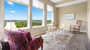 A room with large windows, a view of greenery and water, a wooden rocking chair, a white chair, a table with a lamp, and a patterned rug. - It's a Good Life | Stunning Lake Views! (Branson)