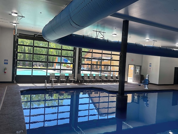 An indoor swimming pool with clear glass walls, a ceiling with a wave-like design, and a view of an outdoor pool area.