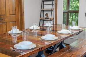 Mesa de jantar posta com pratos brancos, tigelas e copos de vidro transparente, com um banco de madeira e uma escada ao fundo.