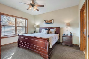 A bedroom with a wooden bed, a ceiling fan, a nightstand, a lamp, and a window with blinds.