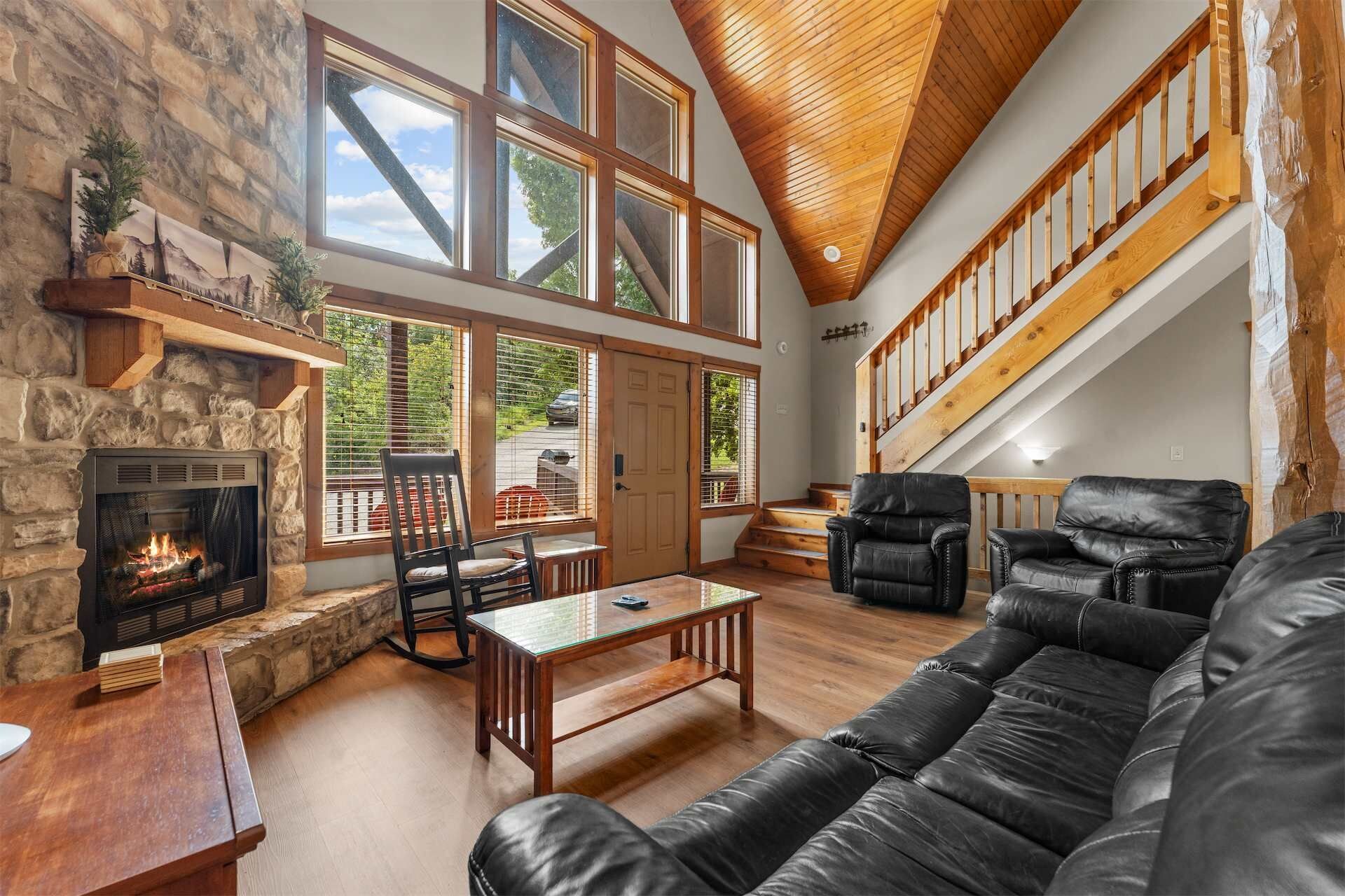 A spacious living room with a stone fireplace, leather furniture, large windows, and a wooden staircase.