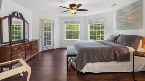 A bedroom with a large bed, a ceiling fan, a wooden dresser, and a window with blinds.