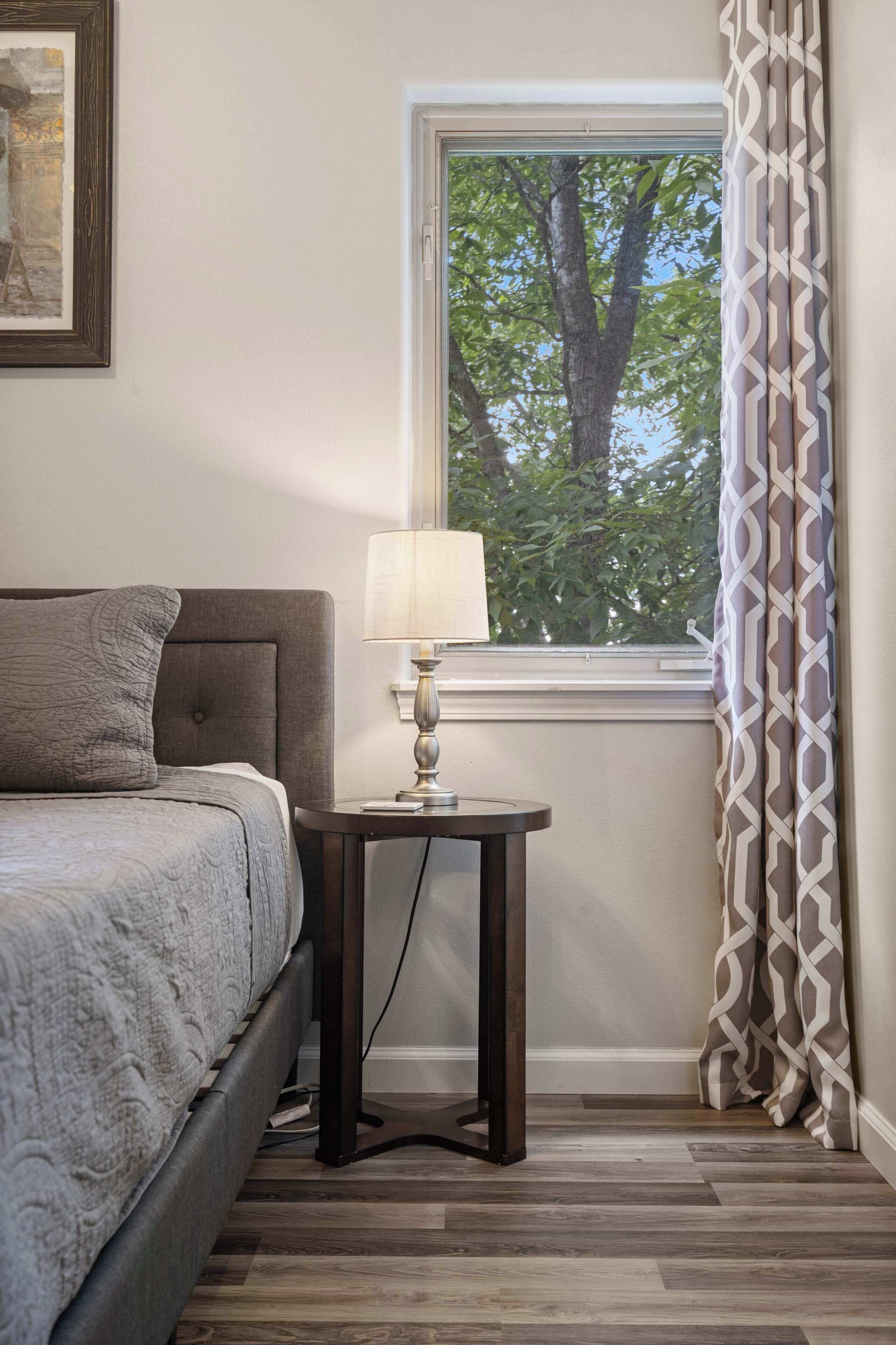 A bedroom with a bed, bedside table, lamp, window with curtains, and a framed picture on the wall.