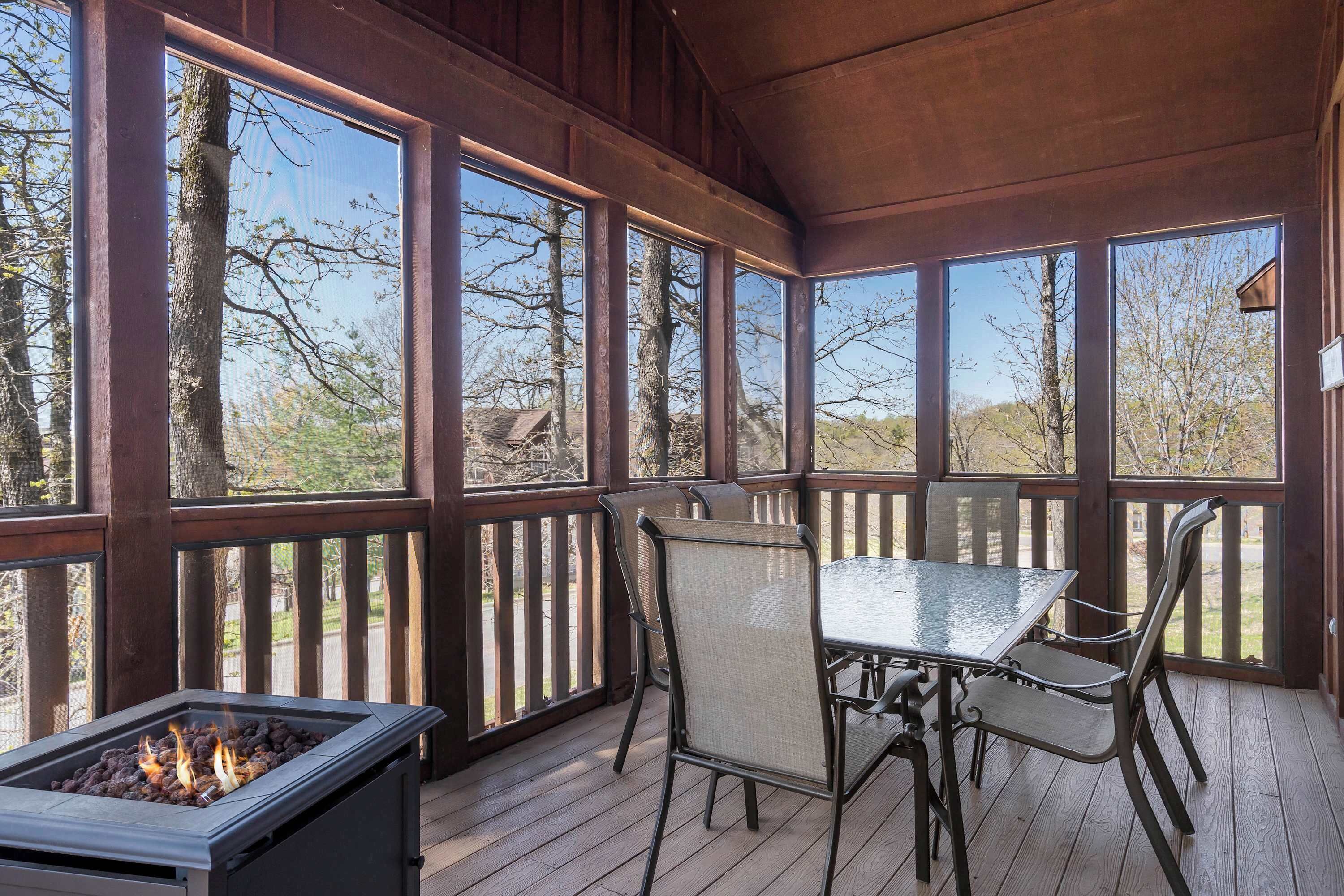A covered outdoor deck with a glass-top table and chairs, a fire feature, and a view of trees and a road.