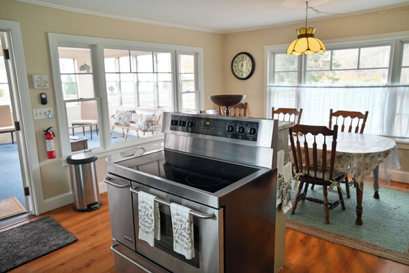 A kitchen with a stainless steel stove, wooden chairs, a dining table with a floral tablecloth, and a clock on the wall. - Charming Rustic Farmhouse. Minutes to Bristol Mountain, Finger Lakes, Wineries. (Naples)