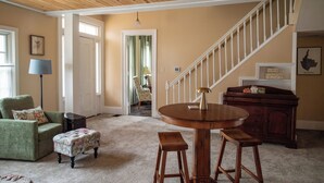 Un vaste salon doté d’un haut plafond, une table ronde en bois avec des tabourets, un fauteuil vert, un petit pouf et un escalier menant à une autre pièce.