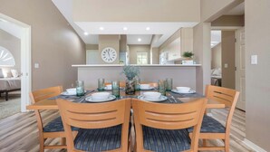 Un comedor con una mesa redonda dispuesta para cuatro personas, sillas de madera con cojines azules, un jarrón con flores y un reloj en la pared.