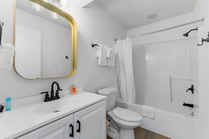 Une salle de bain moderne avec un lavabo, des toilettes et une baignoire blancs, un grand miroir avec un cadre doré et un rideau de douche.