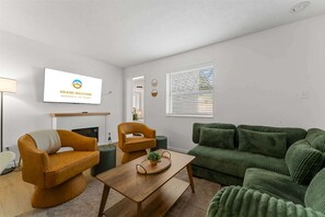 Un salon avec un téléviseur affichant le message « Bienvenue », un canapé, un fauteuil, une table basse et une fenêtre avec des stores.