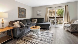 Ein Wohnzimmer mit einer grauen Eckcouch, einem runden Couchtisch, einem gemusterten Teppich und Blick durch eine Glasschiebetür auf einen Außenbereich.