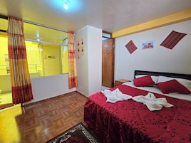 Habitación de hotel con cama, suelo de madera, espejo, armario y ventana con cortinas.