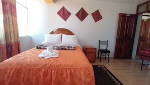 Un lit bien fait, recouvert d’une couvre-lit à motifs floraux, avec une tête de lit en bois et un ensemble de trois coussins décoratifs. Une chaise et une petite table de chevet en bois se trouvent à côté du lit.