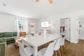 Quarto de hotel moderno com uma mesa de jantar branca, cadeiras brancas, um sofá verde, piso de madeira e uma televisão exibindo 'GRAND WELCOME'.