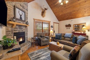 Un salon chaleureux avec une cheminée en pierre, une tête de cerf accrochée au mur, un plafond en bois et des sièges confortables.
