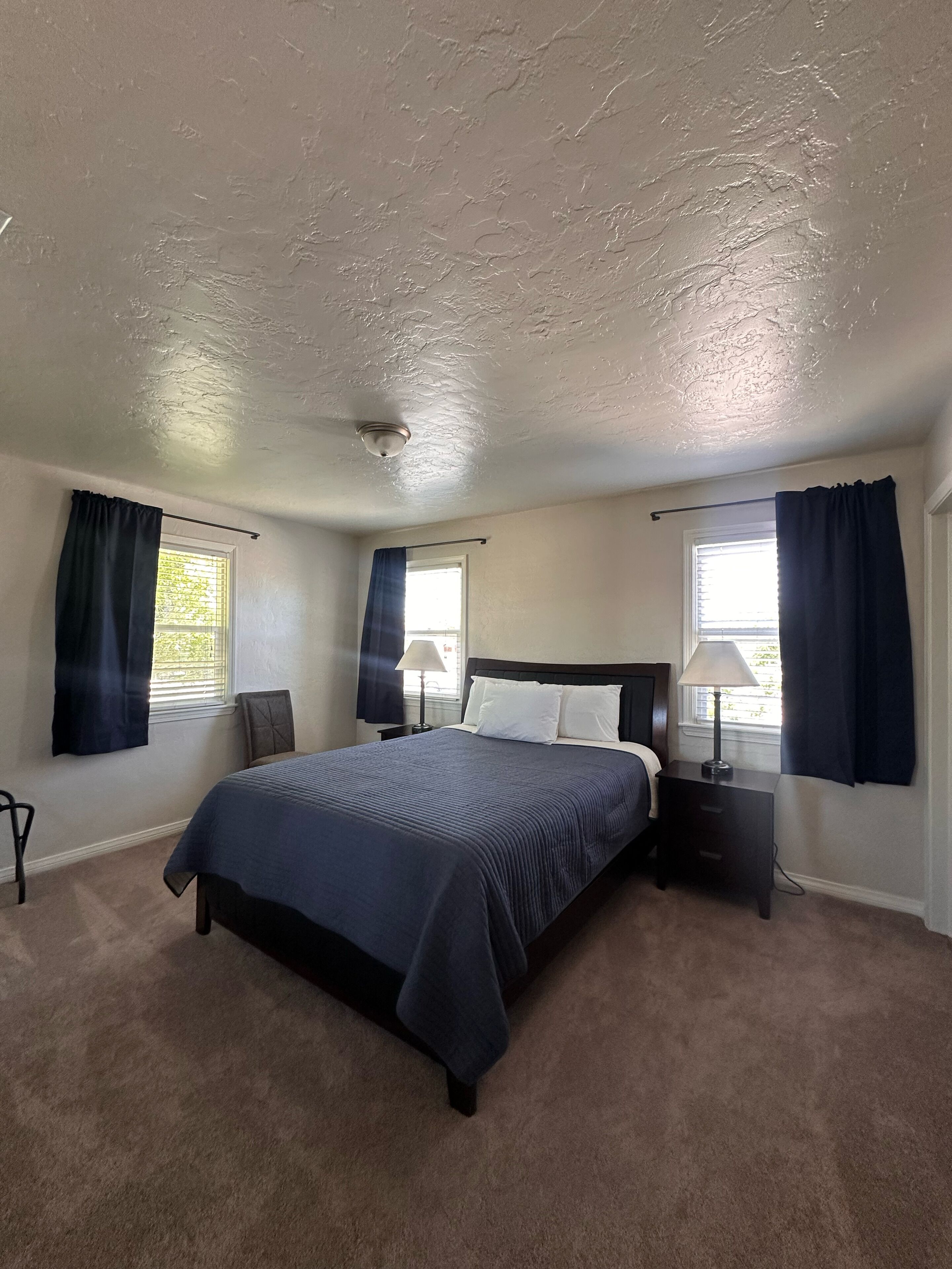 Une chambre à coucher comprenant un lit, deux tables de chevet, une chaise et deux fenêtres avec des rideaux.
