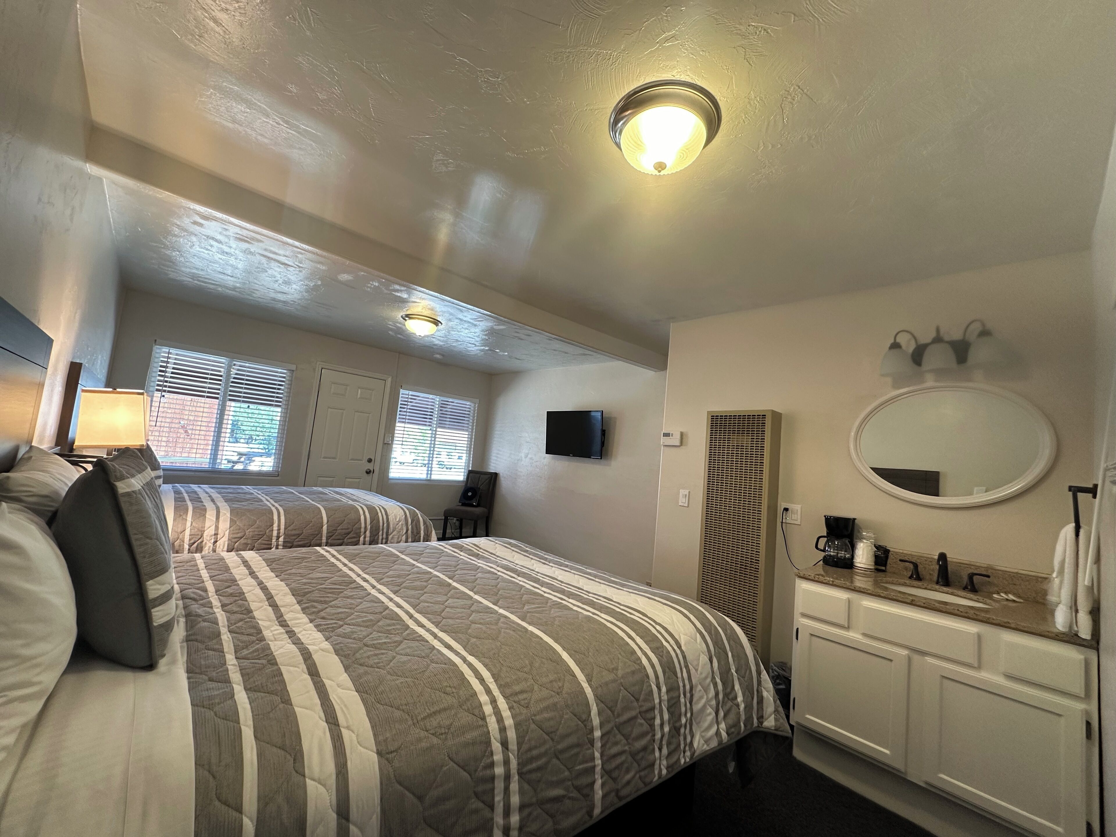 A bedroom with two beds, a TV, a mirror, a sink, and a window with blinds.