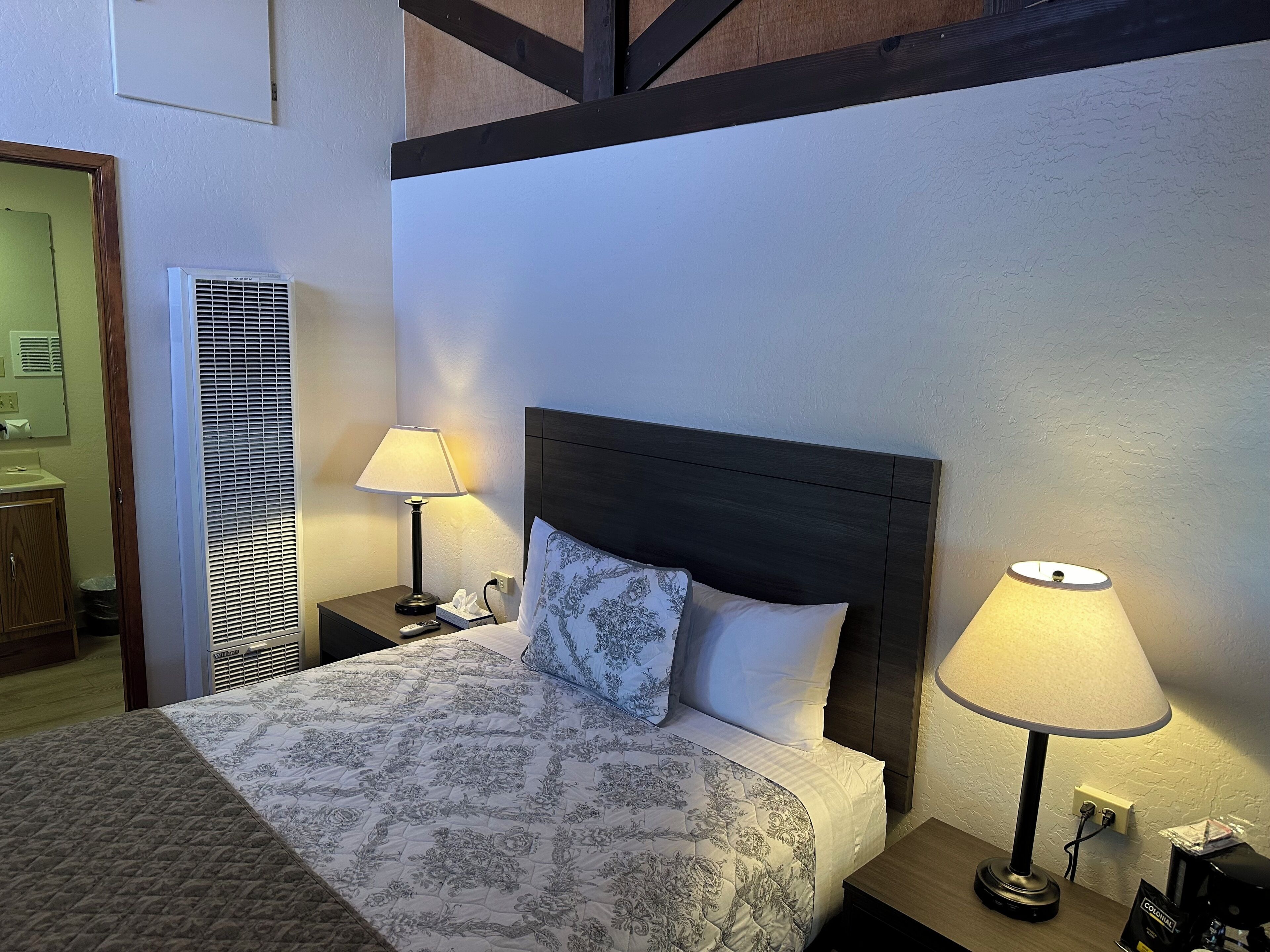Ein Hotelzimmer mit einem Bett, zwei Nachttischlampen, einem Nachttisch, einem hölzernen Kopfteil und einer Wand-Klimaanlage.