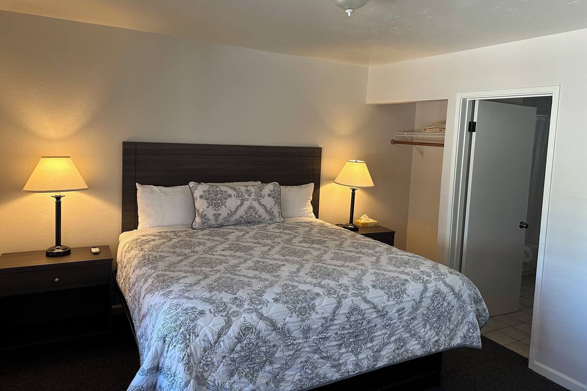 Una cama bien tendida con una colcha estampada, dos lámparas de noche, un cabecero de madera oscura y un ventilador de techo.