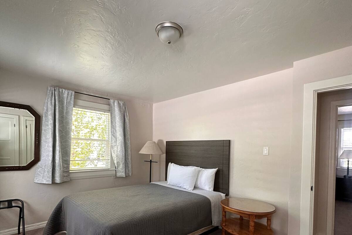 Un dormitorio con una cama, una mesita de noche, una lámpara, una silla y un ventanal con cortinas.