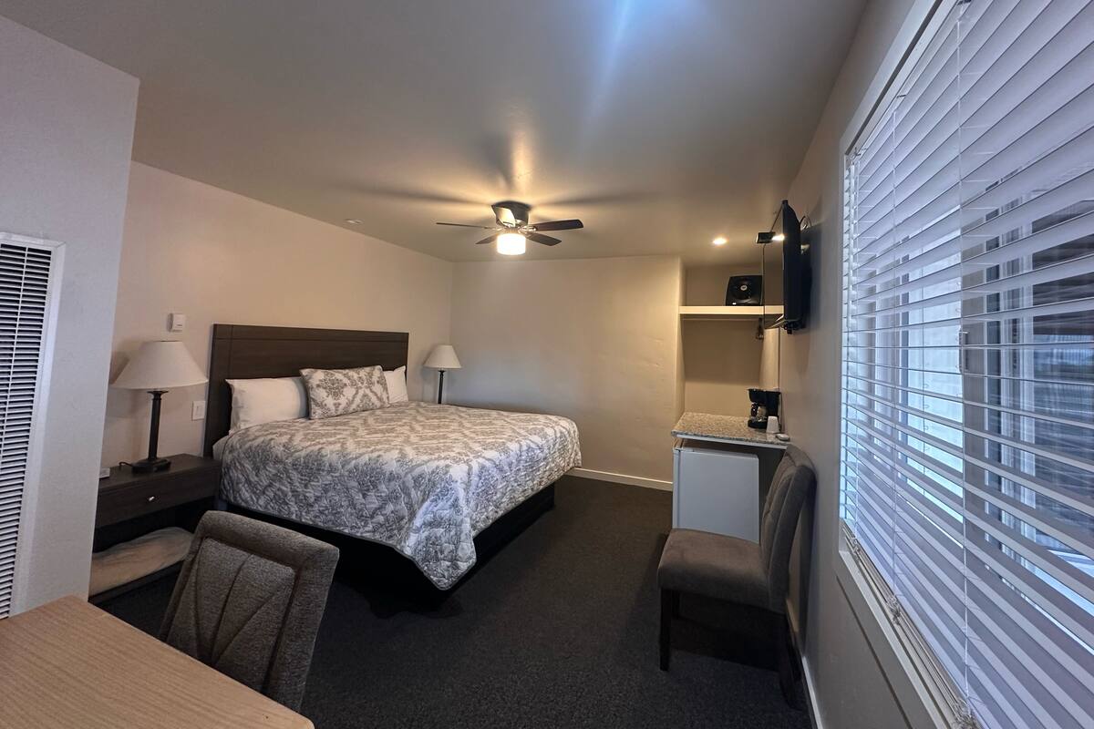 Habitación de hotel con cama, escritorio, silla, ventilador de techo, televisor y ventana con persianas.