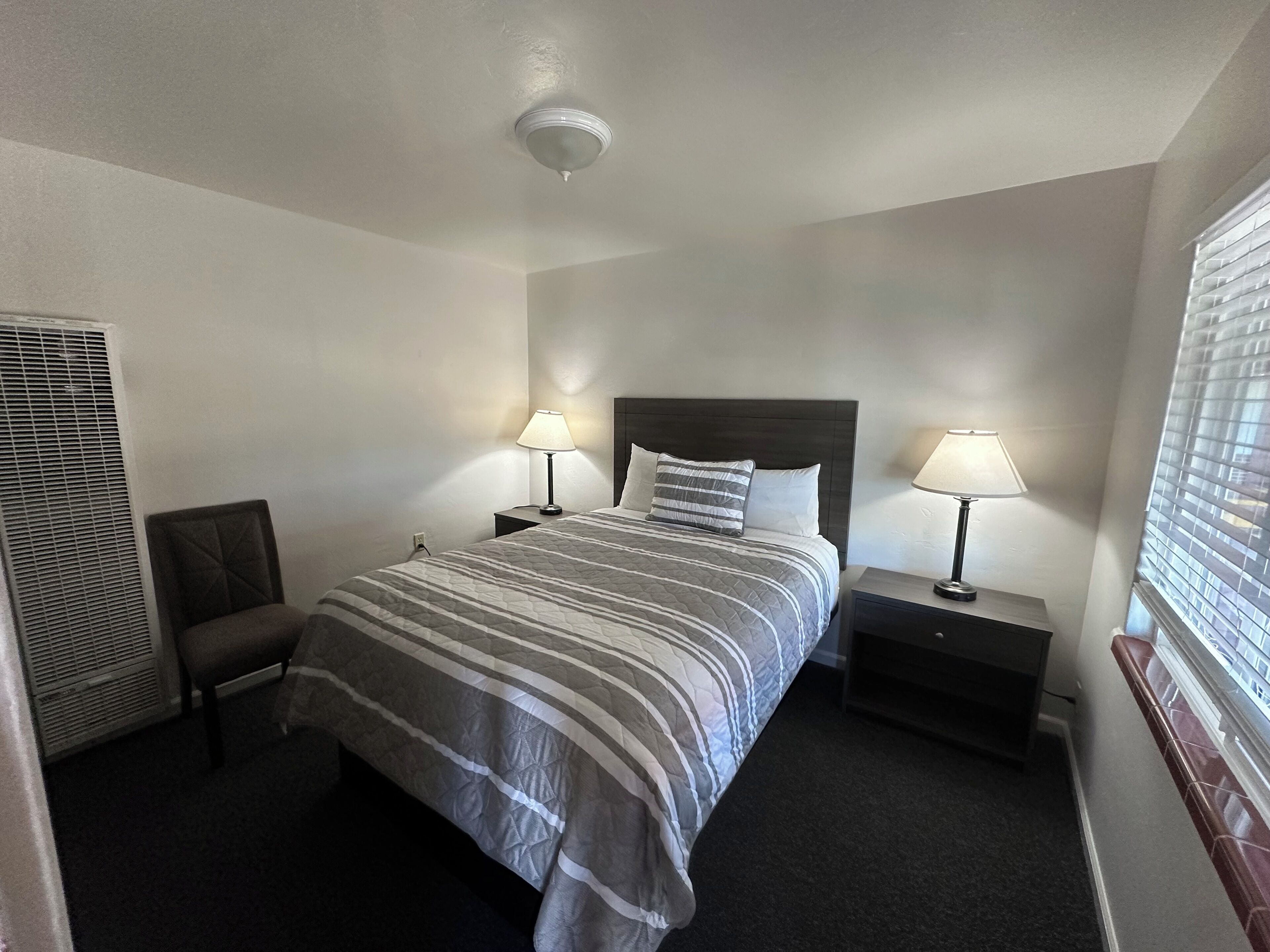A hotel room with a bed, two bedside tables with lamps, a chair, a window with blinds, and a ceiling light.