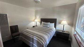 A hotel room with a bed, two bedside tables with lamps, a chair, a window with blinds, and a ceiling light.