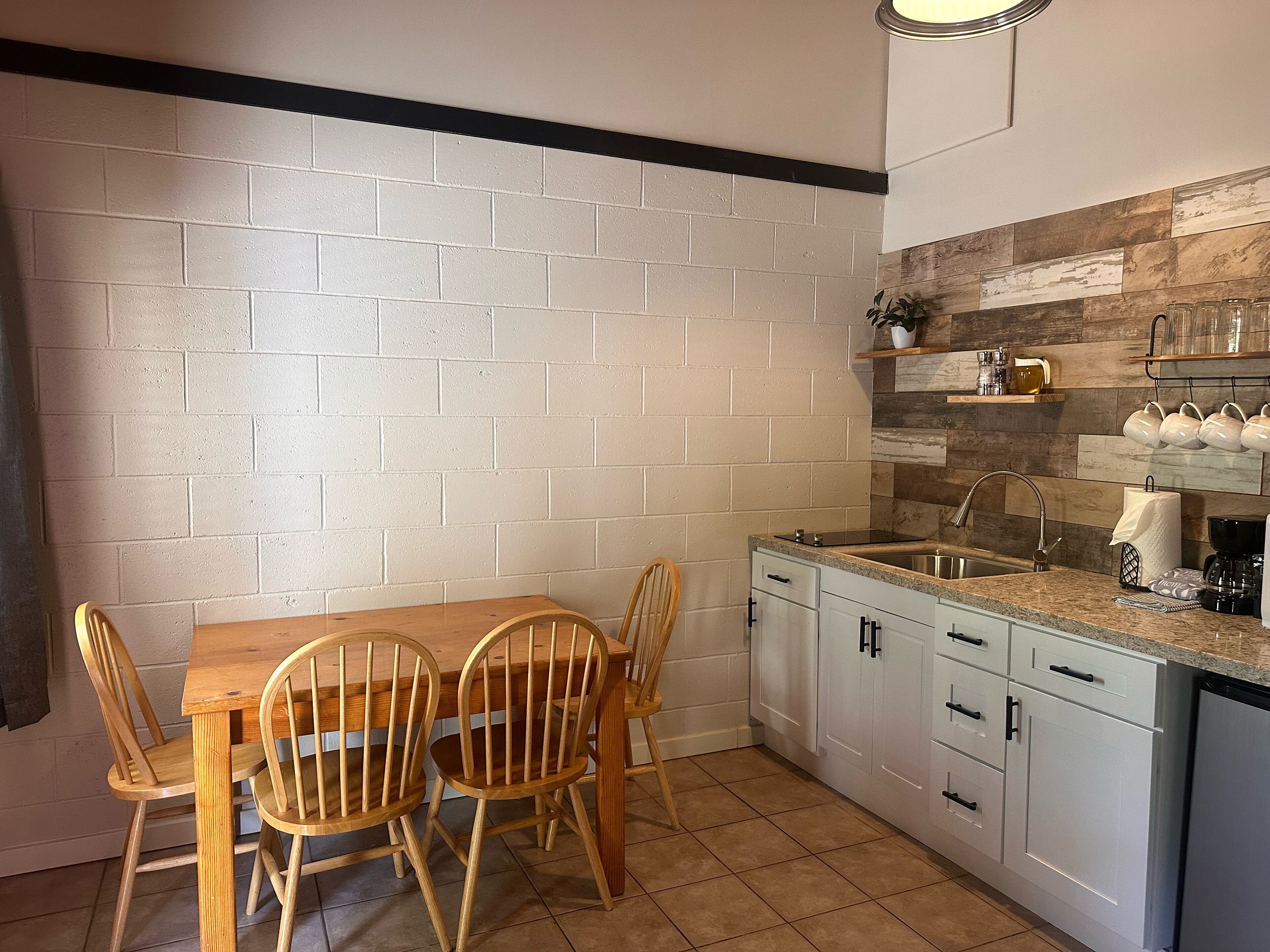 A compact kitchen with white brick walls, a wooden dining table, and light-colored cabinets.