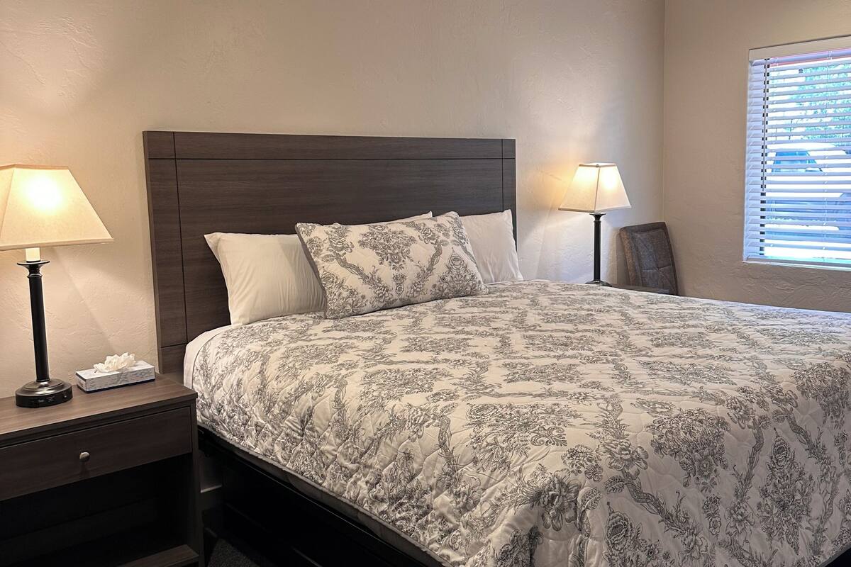 Una cama bien hecha con colcha de estampado floral, dos lámparas de noche, una mesita de noche de madera, un banco y un ventilador de techo.