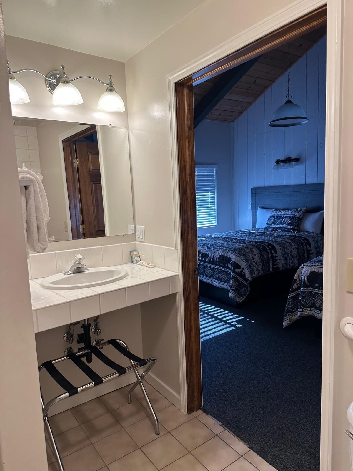 Une salle de bain avec un lavabo blanc, un miroir et un porte-serviettes. Une porte donne sur une chambre à coucher équipée d’un lit et d’un luminaire suspendu.