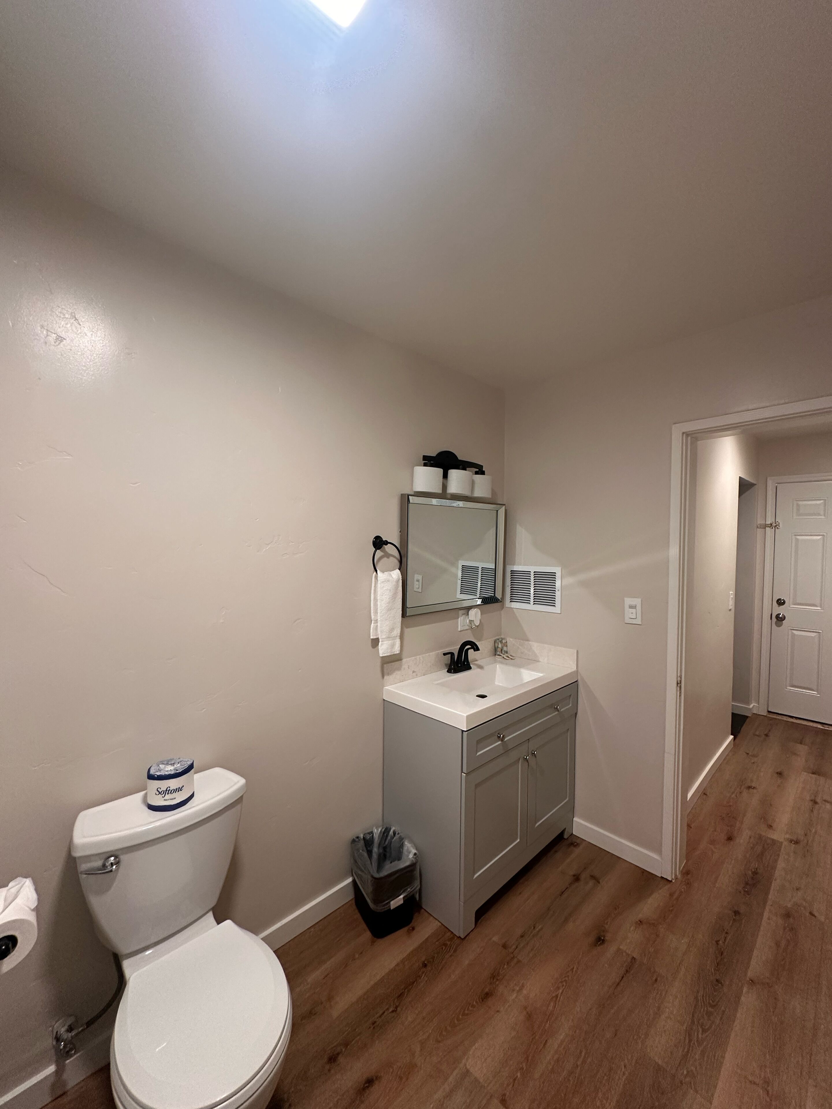 A bathroom with a toilet, sink, and mirror. There is a trash can and a towel hanging on the wall.