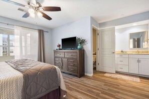 Un dormitorio con cama, televisor sobre un aparador, ventilador de techo y baño con espejo y lavabo.