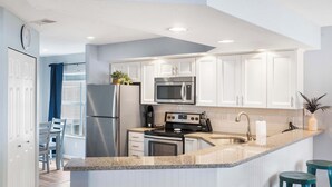 A modern kitchen with white cabinets, a stainless steel refrigerator, a microwave, an oven, and a sink. There are two stools and a potted plant. - Brookside Haven – Gorgeous, Private Community! (Branson)