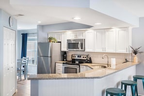 A modern kitchen with white cabinets, a stainless steel refrigerator, a microwave, an oven, and a sink. There are two stools and a potted plant.