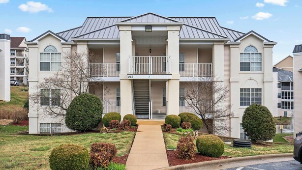 A two-story apartment building with a central entrance, a balcony on the second floor, and a number "215" sign. - Brookside Haven – Gorgeous, Private Community! (Branson)