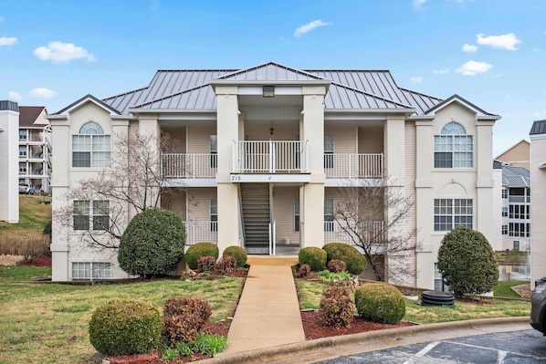 A two-story apartment building with a central entrance, a balcony on the second floor, and a number "215" sign.