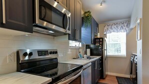 A modern kitchen with stainless steel appliances, a microwave, oven, and dishwasher. There is a window with a floral curtain and a small plant on the wall.